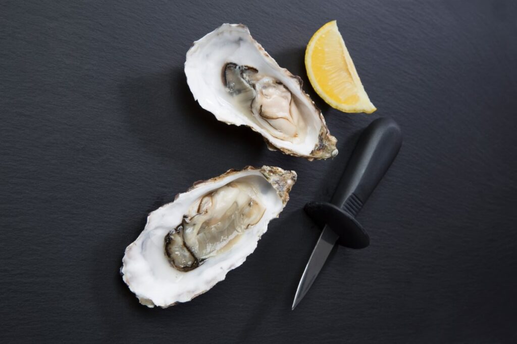 Open oysters, an oyster knife and a slice of lemon arranged on a black background