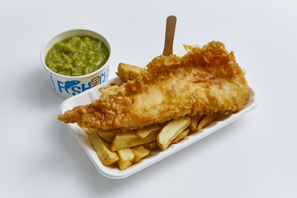 Fish and chips on a bio box tray with a wooden fork and a pot of mushy peas